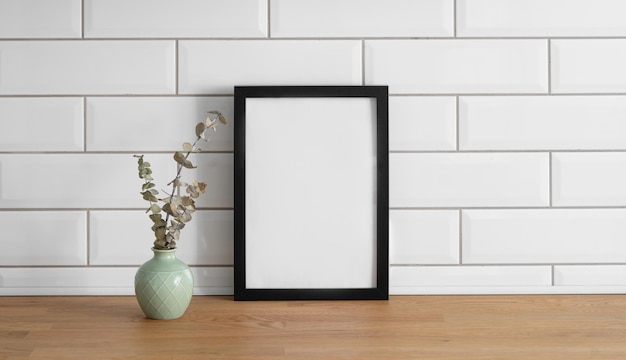 A frame with a blank canvas against a white tile wall and on a wooden tabletop with vase