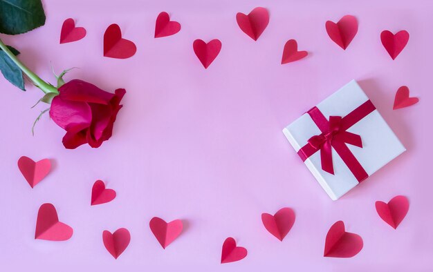 Photo frame with fresh red rose and gift box and paper hearts and pink paper background