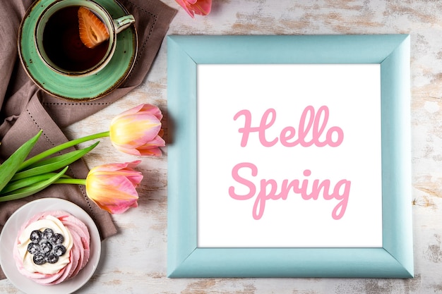 Frame with the inscription Hello Spring, pink tulips, cake and a cap of tea on a white wooden background. High quality photo