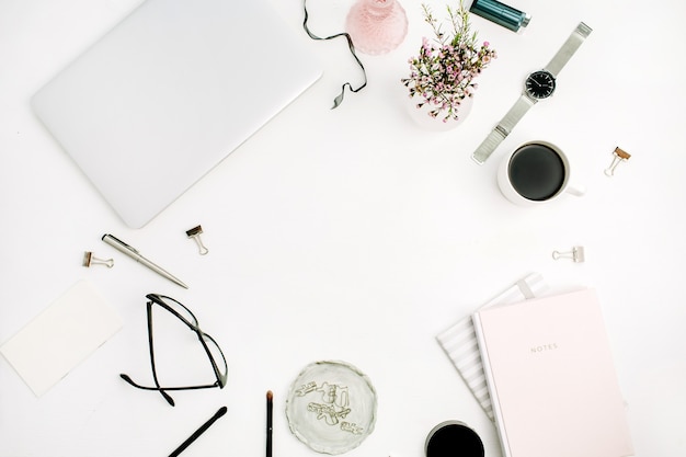 Frame of woman modern home office space with laptop, pastel pink notebook, glasses, coffee cup, wild flowers and accessories on white desk. Flat lay, top view