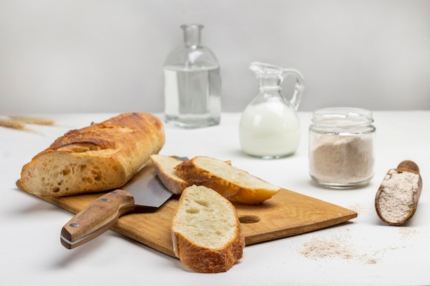 Franse stokbrood plakjes en een mes op een snijplank. Meel in een schep en glazen pot. Melk en water in glazen flessen. Witte achtergrond.