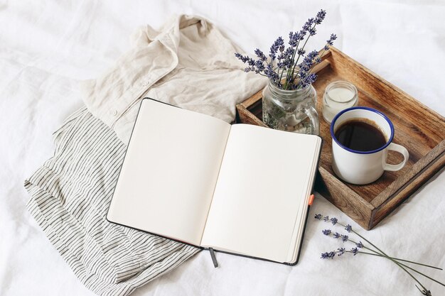 Franse zomer stilleven Vrouwelijke levensstijl samenstelling Kopje koffie lavendel bloemen boeket kaars op houten dienblad Linnen overhemd en broek op bed Lege notebook mockup scène Bovenaanzicht