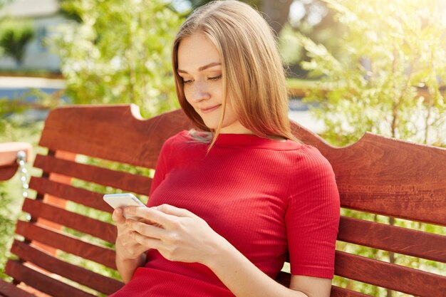 Freckled girl with straight hairstyle wearing red dress sitting in park at bench using her cell phone surfing social networks