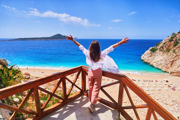 Free carefree traveler girl stands alone with open arms on stairs against backdrop of bay with the turquoise sea in Kas Turkey