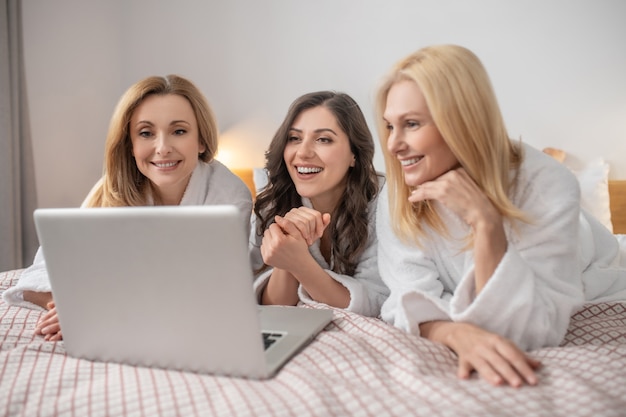 Free time. Good looking cheerful middle aged women in white coats lying on bed looking at laptop communicating