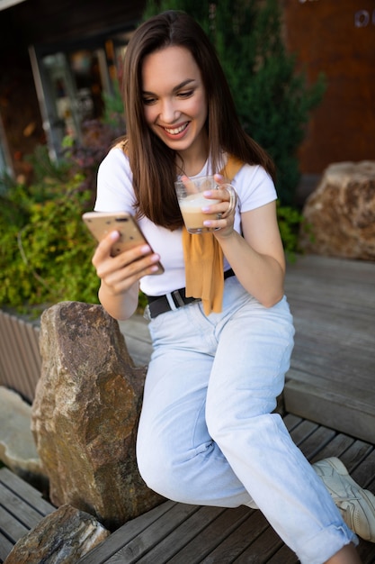 サマーパークのベンチに座ってコーヒーを飲み、スマートフォンでチャットする無料の若い女性。