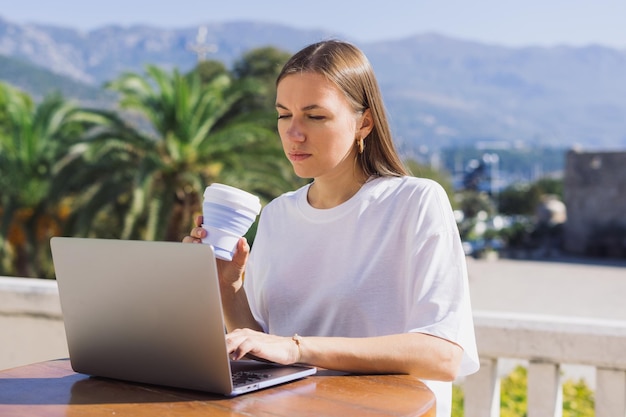 Свобода от работодателя Работа из любой точки мира Девушка пьет кофе и пользуется ноутбуком в летнем кафе