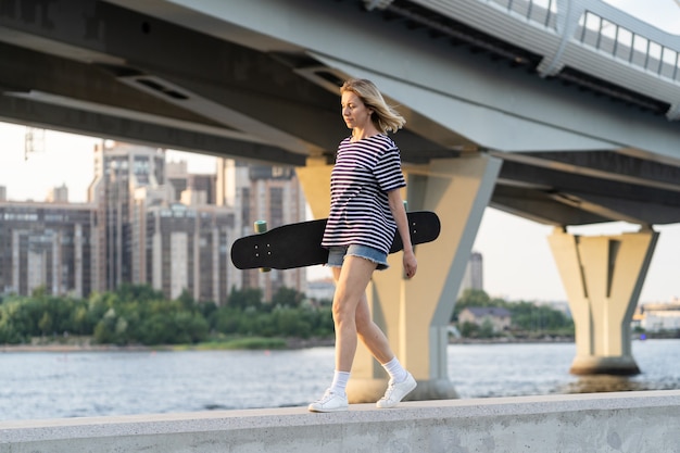 Freedom lifestyle woman of middle age walk near river hold longboard over big city view at sunset