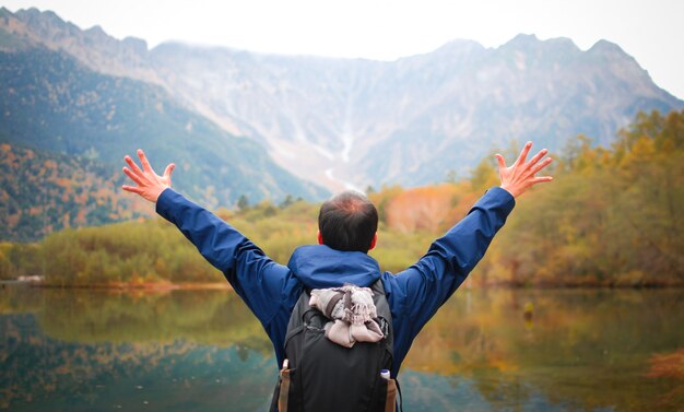 Uomo di libertà con le mani in alto sul lato del lago contro la foresta e le montagne naturali di autunno