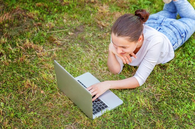 Freelance bedrijfsconcept. Jonge vrouw liggend op groen gras gazon in stadspark werken op laptop pc-computer. Lifestyle authentiek openhartig studentenmeisje dat buitenshuis studeert. Mobiel kantoor