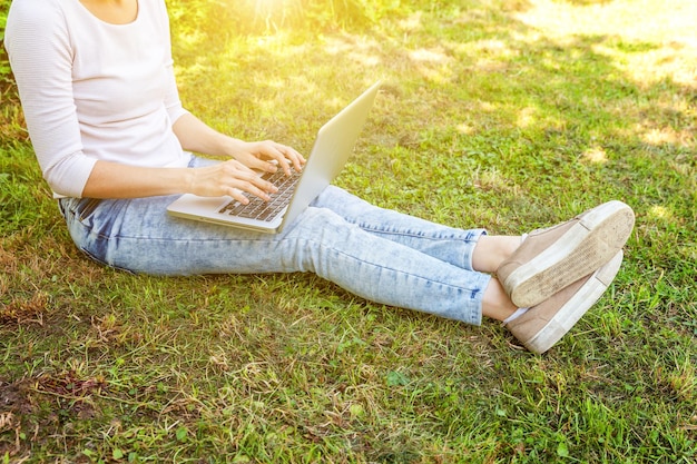 Freelance business concept. Woman legs on green grass lawn in city park, hands working on laptop pc computer. Lifestyle authentic candid student girl studying outdoors. Mobile Office