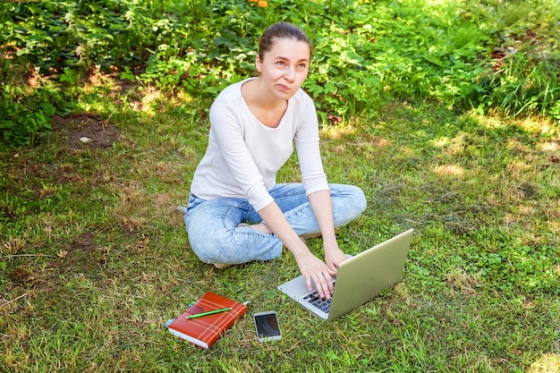 Freelance business concept. Young woman sitting on green grass lawn in city park working on laptop pc computer. Lifestyle authentic candid student girl studying outdoors. Mobile Office