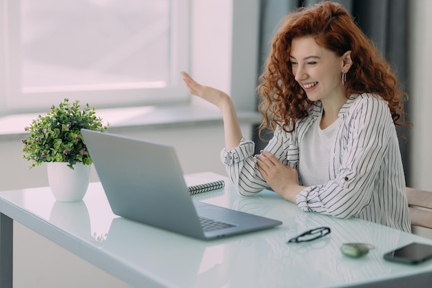 Photo freelance business woman with ginger hair sits in front of laptop computer communicates with colleagues via video conference sits at desktop drinks coffee has happy expression distance job