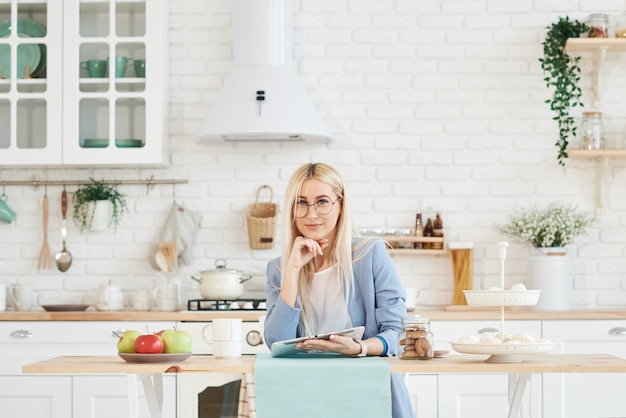 Freelance concept. Beautiful business woman in casual clothes and glasses is examining documents and smiling while working with a laptop in kitchen.