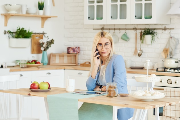 Freelance concept. Beautiful business woman in casual clothes and glasses is examining documents and smiling while working with a laptop in kitchen