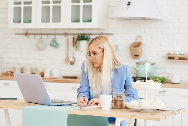 Freelance concept. de mooie bedrijfsvrouw in vrijetijdskleding en glazen onderzoekt documenten en glimlacht terwijl het werken met laptop in keuken