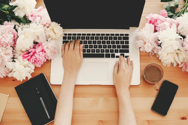 Photo freelance concept hands working on stylish laptop with empty screen coffee cup notebookphone pink and white peonies on wooden table flat lay with space for text international womens day