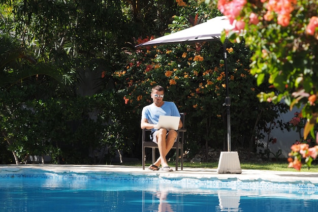 フリーランスのコンセプト。暖かい気候のラップトップを持った男がプールの近くに座って働いています。庭の周り。