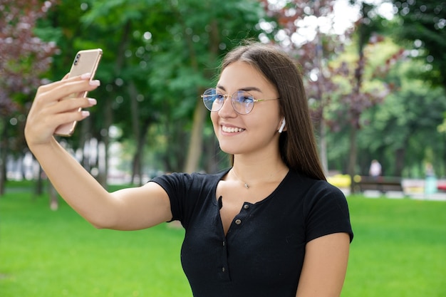 Freelance, fresh air, business, technology, and Internet concept â Optimistic European woman wearing eyeglasses and wireless headphones conducts a video communication session using the phone.