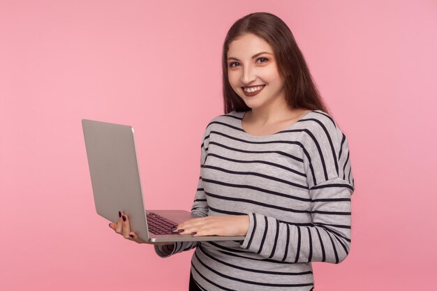 フリーランスの仕事、オンライン学習。ラップトップを保持し、カメラに微笑んで、コンピューターで作業している縞模様のスウェットシャツで幸せなフレンドリーな若い女性の肖像画。ピンクの背景に分離された屋内スタジオショット
