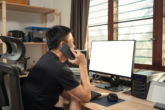 Freelance jonge aziatische man belt met mobiele telefoon en leeg display, opbergdoos aan houten bureau aan raamzijde in huis