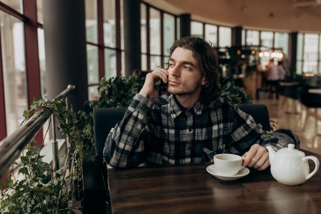 Freelance man kijkt peinzend uit het raam en praat aan de telefoon terwijl hij op afstand werkt in een gezellig café