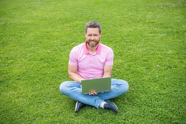 フリーランスの男性がブログをオンラインで投稿し、芝生の上に座っています。オンラインで書く。ブログにラップトップを使用しているビジネスマン。ソーシャルネットワークとウェブログ。現代のビジネスコンピュータ通信。笑顔の男はメールをチェックします。