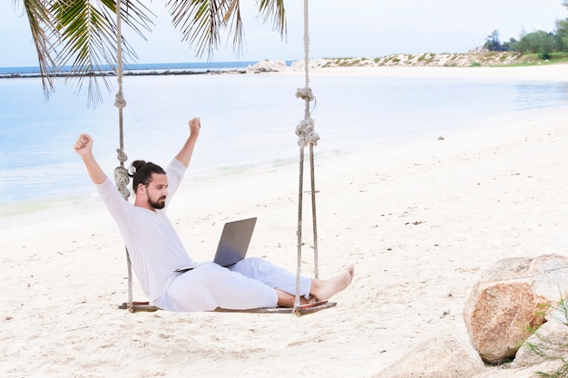 Внештатный мужчина в белом сидит на качелях на пляже с ноутбуком