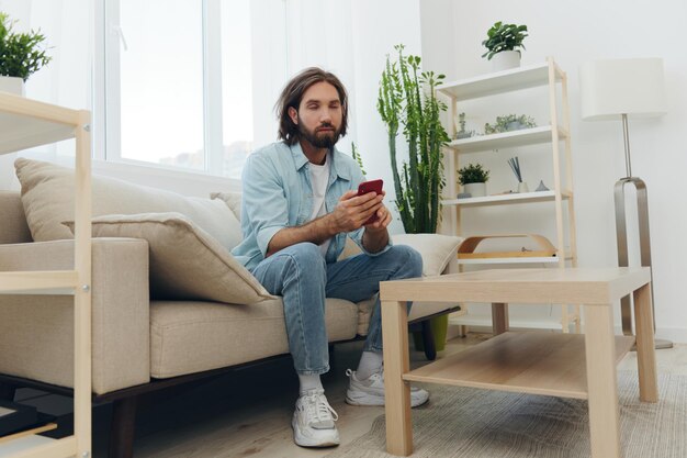 a freelance man in a white tshirt blue jeans and shirt sits on the couch with his phone at home on his day off and plays games high quality photo
