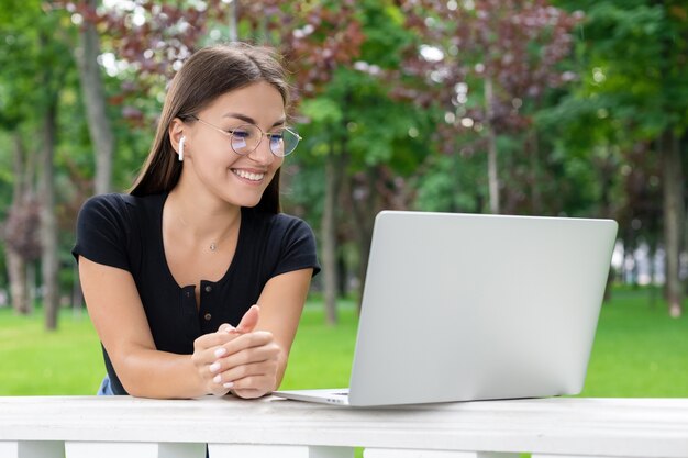 Freelance, onderwijs, frisse lucht, zaken, technologie en internetconcept - Optimistische Europese vrouw met bril en draadloze koptelefoon die bij zonnig weer naar een laptop in het park kijkt.