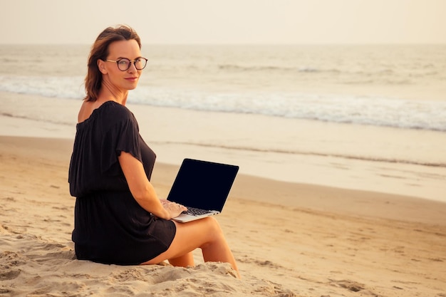 Freelance and remote workbusiness woman in a stylish business summer suit with laptop sitting in the tropical ocean coast palm sandfemale student with glasses online training education abroad