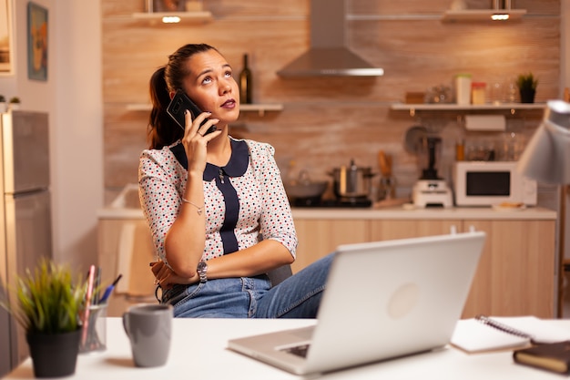 Donna freelance che ha una conversazione telefonica con un cliente a tarda notte dalla cucina di casa. dipendente che utilizza la tecnologia moderna a mezzanotte facendo gli straordinari per lavoro, affari, carriera, rete, stile di vita.