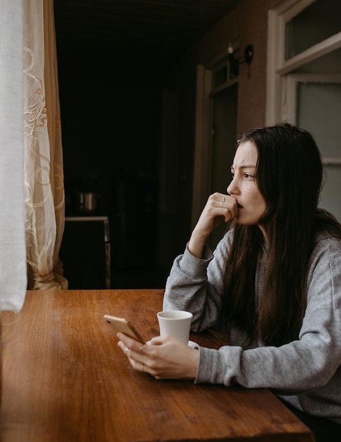 写真 コーヒーと携帯電話を持ったフリーランス女性が家で深く考えています