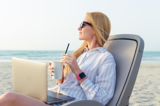 A freelance woman works at a laptop and drinks a cocktail by the sea