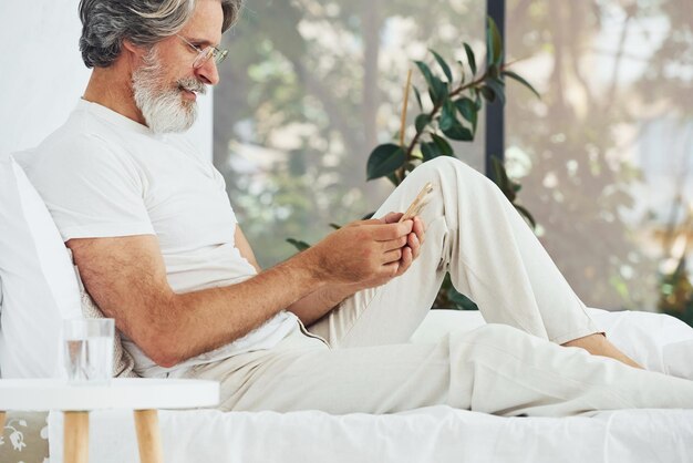 Внештатный работник на кровати с ноутбуком Старший стильный современный мужчина с седыми волосами и бородой в помещении
