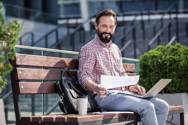 Freelance worker. Cheerful bearded man using his laptop while sitting on the bench