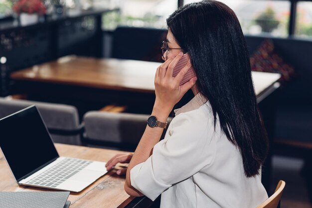 ラップトップ コンピューターを持つフリーランスの働く女性、コーヒー ショップで顧客と話しているスマート フォンを呼び出す彼