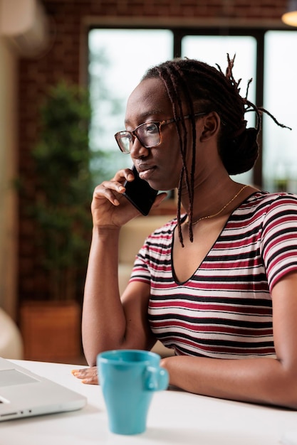 Freelancer answering mobile phone call, drinking tea in home office. Busy african american remote worker chatting on smartphone, sitting at desk, employee telephone comuunication