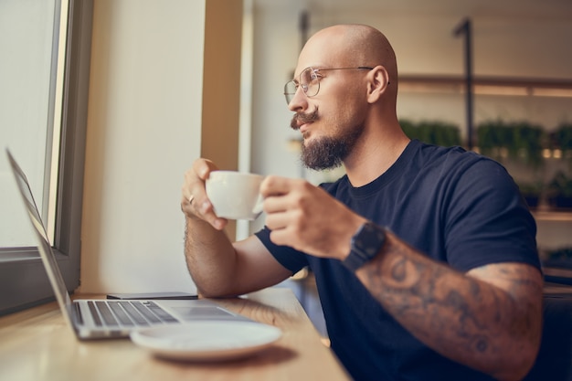 Freelancer bald man in glasses with mustache and beard works in cafe uses laptop while drinks coffe