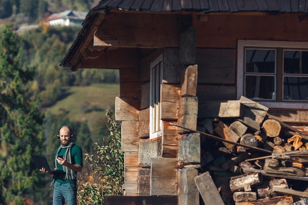 Freelancer bearded man is standing near log house in mountains with laptop and talking on phone.