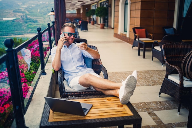Концепция фрилансера Успешный молодой бизнесмен, работающий на ноутбуке, сидит на террасе, пьет кофе и разговаривает по телефону