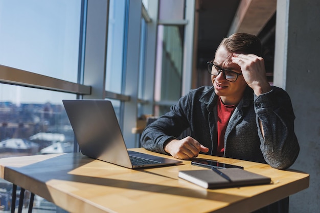 A freelancer in glasses looks into a laptop and he is angry while sitting at a table with a laptop and a notepad in a cafe during the day Unlucky day Working remotely from the office