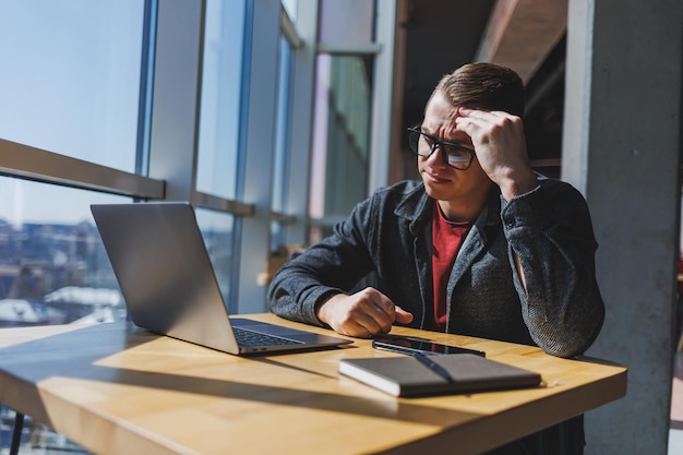 A freelancer in glasses looks into a laptop and he is angry while sitting at a table with a laptop and a notepad in a cafe during the day Unlucky day Working remotely from the office