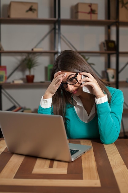 Freelancer is working with something and sitting at the table with laptop and speaking on phone