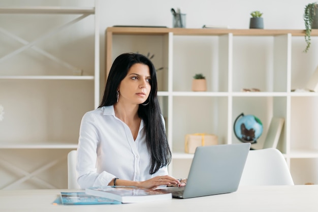 Freelancer levensstijl Externe baan Virtueel kantoor Doordachte serieuze zakenvrouw die op laptop werkt en wegkijkt op een lichte werkplek