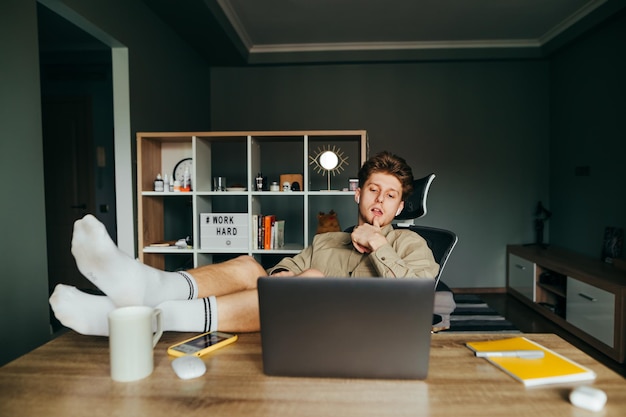 freelancer ligt thuis in een stoel op het werk met zijn benen op tafel en gebruikt een laptop