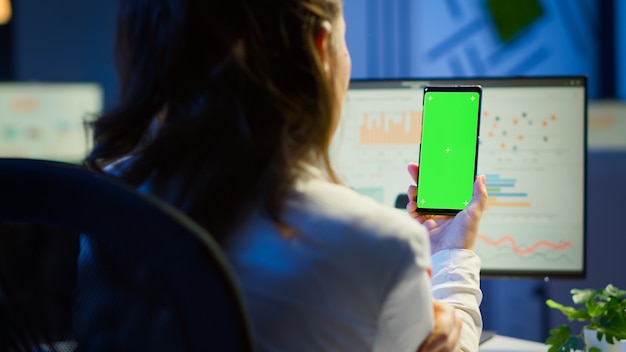 Foto libero professionista che guarda il telefono cellulare con schermo verde durante la riunione video seduto alla scrivania in ufficio a tarda notte. donna d'affari che guarda monitor desktop con mockup verde, chroma key