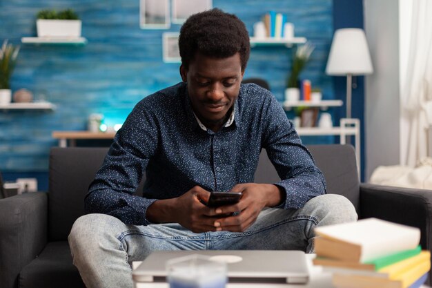 Человек-фрилансер просматривает социальные сети с помощью смартфона в свободное время в гостиной. черный подросток ищет информацию в интернете, болтает с виртуальными друзьями, отправляя текстовые сообщения