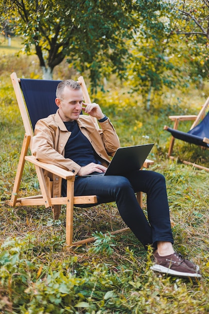 Freelancer man met laptop aan het werk in de tuin zittend in een houten stoel buitenshuis Een jonge blogger werkt op een computer in een openbaar park Werkplek op afstand werken