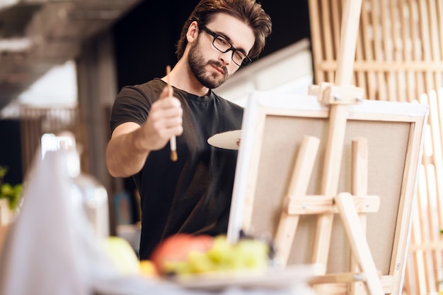 Freelancer man painting with brush standing behind easel.